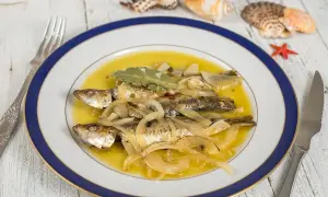 Tres sardinas en escabeche, cubiertas de cebolla y laurel, nadan en un líquido dorado. El plato está decorado con conchas marinas.