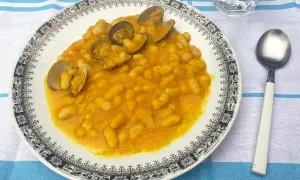 Plato de habichuelas con almejas, servido en un plato blanco con borde decorado. Acompañado de cuchara y vaso de agua.