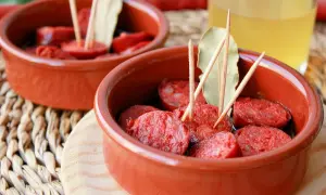 Dos cuencos de barro con rodajas de chorizo a la sidra, adornados con palillos y hojas de laurel.