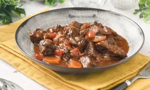 Plato humeante de carne al toro, tiernos trozos de carne en salsa oscura con zanahorias, servido en un cuenco gris sobre servilleta amarilla.