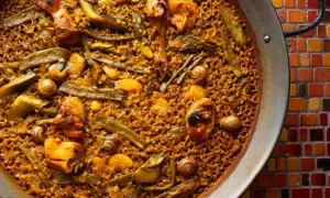 Paella de pollo con trozos de pollo dorado, judías verdes y caracoles, servida en una paellera sobre un mosaico de colores.