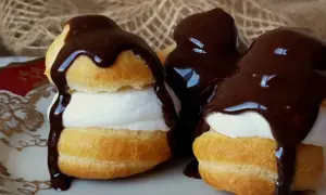 Deliciosos profiteroles de nata cubiertos con chocolate negro derretido, servidos en un plato blanco con detalles dorados.