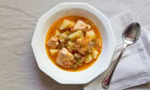 Un plato de marmitako de salmón con patatas y pimientos en una cazuela blanca, acompañado de una cuchara.