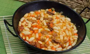 Un plato humeante de alubias con chorizo, zanahoria y patata en una paellera negra, servido sobre una esterilla verde.