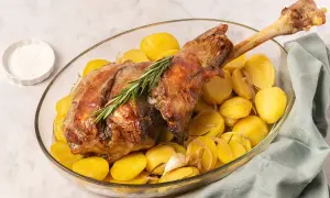 Pierna de cordero al horno dorada y jugosa, servida con patatas asadas, romero y ajo en una fuente de cristal.