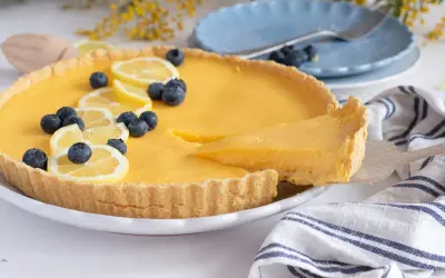 Deliciosa tarta de limón, decorada con rodajas de limón y arándanos. Una porción se sirve con una espátula.
