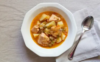 Un plato de marmitako de salmón con patatas y pimientos en una cazuela blanca, acompañado de una cuchara.