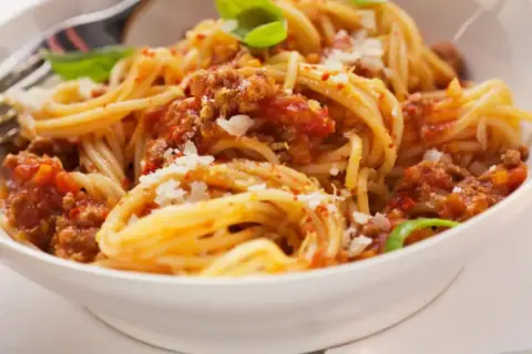 Un plato de espaguetis con carne picada y tomate, espolvoreado con queso rallado y decorado con hojas de albahaca.