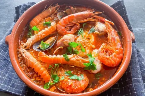Zarzuela de pescado en cazuela de barro, con gambas, cigalas, mejillones y trozos de pescado en salsa roja, decorada con perejil.