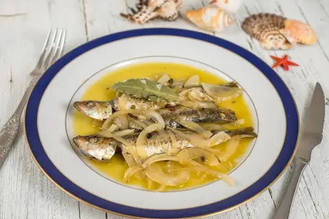 Tres sardinas en escabeche, cubiertas de cebolla y laurel, nadan en un líquido dorado. El plato está decorado con conchas marinas.