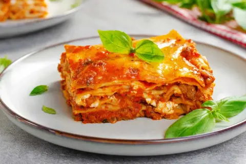 Porción de lasaña de carne con capas de pasta, salsa de tomate y queso, decorada con hojas de albahaca fresca.