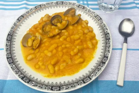 Plato de habichuelas con almejas, servido en un plato blanco con borde decorado. Acompañado de cuchara y vaso de agua.