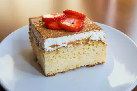Un trozo de tarta tres leches esponjosa, cubierta con crema batida, canela y fresas frescas, servida en un plato blanco.
