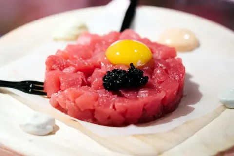 Delicioso tartar de atún fresco, coronado con yema de huevo y caviar negro. Servido con salsas cremosas y un tenedor.