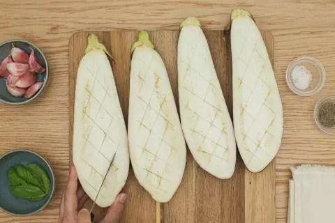 Cuatro mitades de berenjena cortadas en rejilla, listas para ser rellenas. A su lado, ajo, albahaca, sal y pimienta.