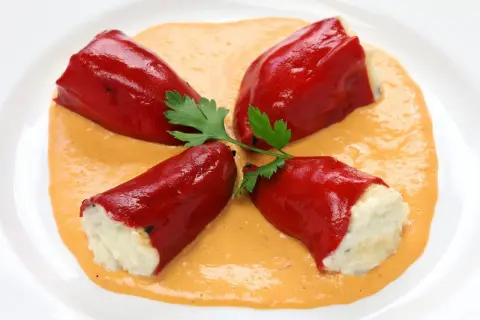 Cuatro pimientos rojos rellenos de bacalao, servidos sobre una cremosa salsa naranja y decorados con perejil.