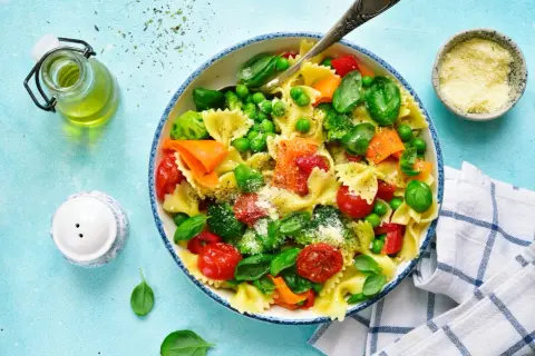 Un plato de pasta con verduras frescas: brócoli, guisantes, tomates cherry y zanahoria, espolvoreado con queso rallado.