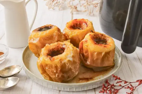 Cuatro manzanas asadas en freidora de aire, doradas y jugosas, servidas en un plato con un jarrito blanco al fondo.