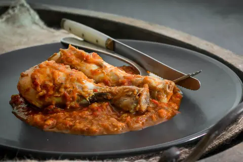 Dos muslos de pollo al chilindrón, servidos en un plato oscuro con cubiertos.