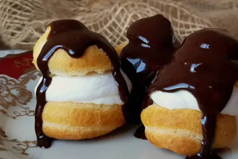 Deliciosos profiteroles de nata cubiertos con chocolate negro derretido, servidos en un plato blanco con detalles dorados.