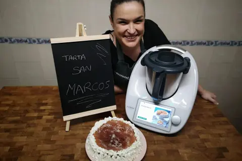 Mujer sonriente junto a una tarta San Marcos y una Thermomix. La pizarra anuncia "Tarta San Marcos".
