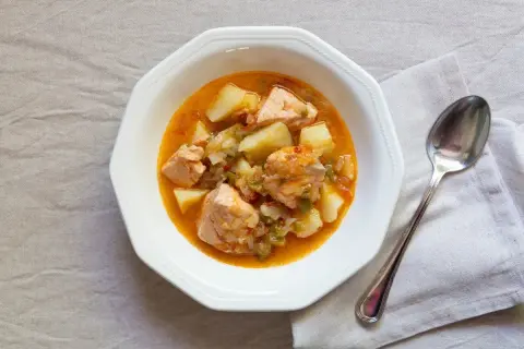 Un plato de marmitako de salmón con patatas y pimientos en una cazuela blanca, acompañado de una cuchara.