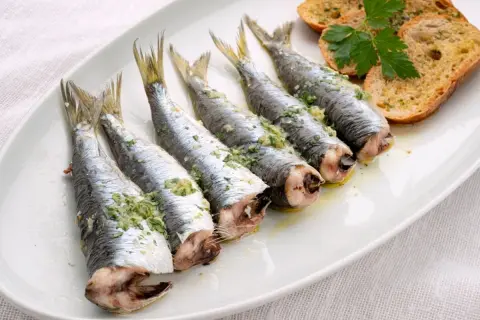 Cinco sardinas al horno, preparadas con hierbas y ajo, servidas en una bandeja blanca con tostadas.