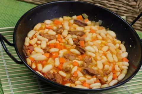 Un plato humeante de alubias con chorizo, zanahoria y patata en una paellera negra, servido sobre una esterilla verde.