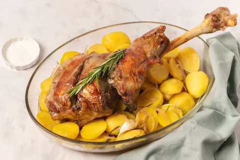 Pierna de cordero al horno dorada y jugosa, servida con patatas asadas, romero y ajo en una fuente de cristal.