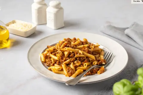 Un plato de macarrones a la boloñesa, con un tenedor listo para disfrutar. Acompañan aceite de oliva, queso rallado y saleros.