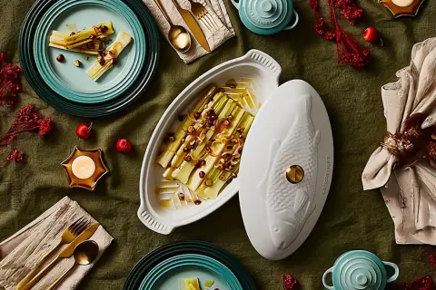 Puerros al horno, tiernos y dorados, servidos en una elegante fuente con tapa en forma de pez. Decoración festiva con velas y frutos rojos.