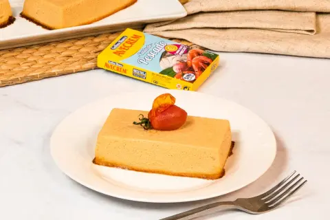 Pastel de merluza servido en plato blanco, decorado con tomate y hierbas. Caja de caldo de pescado Avecrem al fondo.