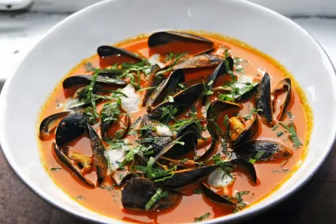 Un tazón blanco rebosante de mejillones al vapor en una salsa roja vibrante, adornado con hierbas frescas picadas.