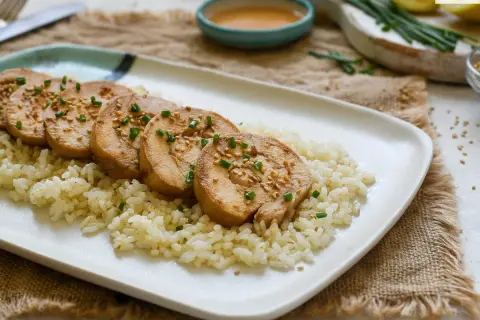 Pechuga de pollo en rodajas, servida sobre arroz blanco y espolvoreada con semillas de sésamo y cebollín.