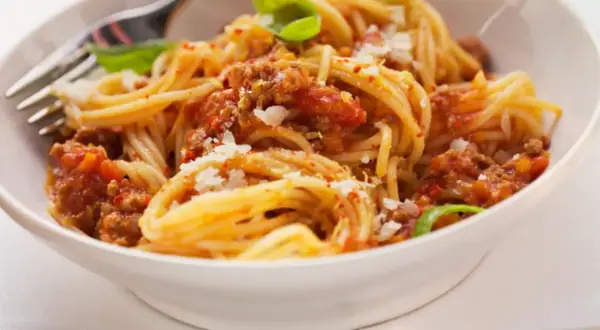 Un plato de espaguetis con carne picada y tomate, espolvoreado con queso rallado y decorado con hojas de albahaca.