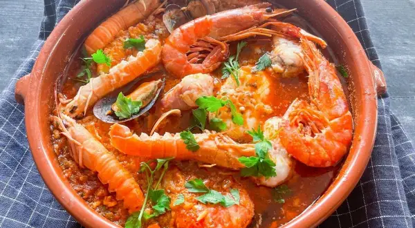 Zarzuela de pescado en cazuela de barro, con gambas, cigalas, mejillones y trozos de pescado en salsa roja, decorada con perejil.