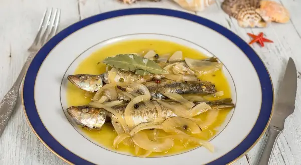 Tres sardinas en escabeche, cubiertas de cebolla y laurel, nadan en un líquido dorado. El plato está decorado con conchas marinas.