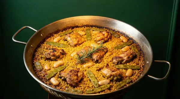 Paella valenciana recién hecha, con trozos de pollo dorado, judías verdes y garrofón, coronada con una ramita de romero.