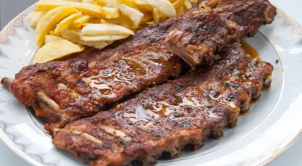 Costillas de cerdo jugosas y doradas, cubiertas de salsa brillante, acompañadas de papas fritas crujientes en un plato blanco.