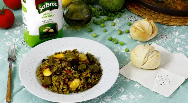 Un plato de paella de verduras, con arroz Sabroz, tomates, alcachofas, guisantes y panecillos.