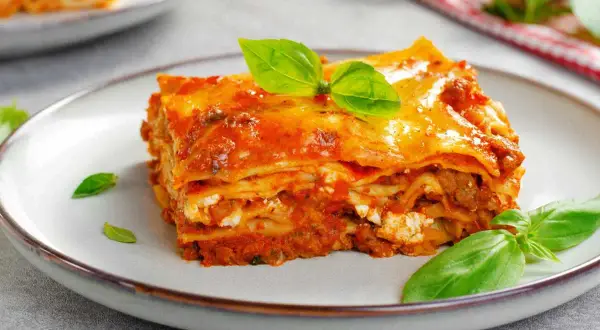 Porción de lasaña de carne con capas de pasta, salsa de tomate y queso, decorada con hojas de albahaca fresca.