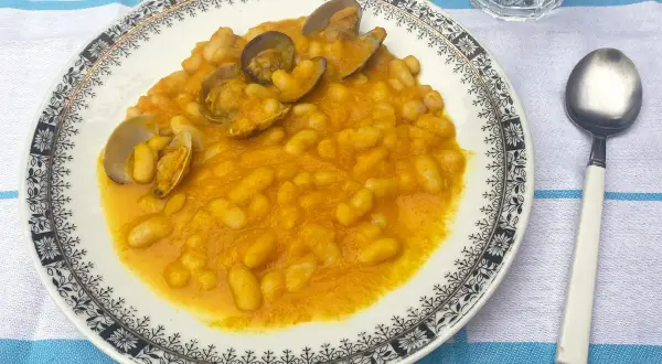 Plato de habichuelas con almejas, servido en un plato blanco con borde decorado. Acompañado de cuchara y vaso de agua.