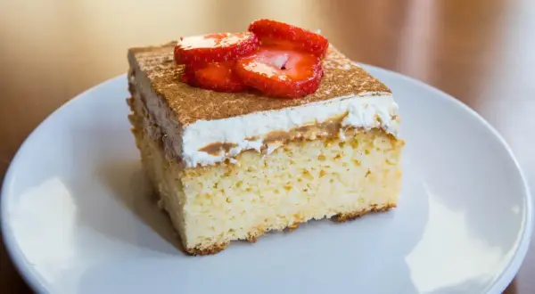 Un trozo de tarta tres leches esponjosa, cubierta con crema batida, canela y fresas frescas, servida en un plato blanco.