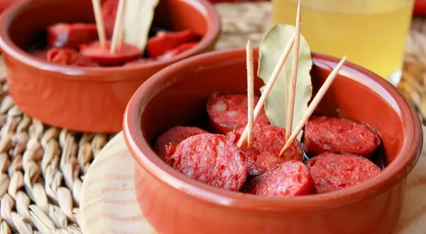 Dos cuencos de barro con rodajas de chorizo a la sidra, adornados con palillos y hojas de laurel.