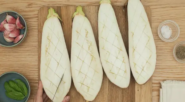 Cuatro mitades de berenjena cortadas en rejilla, listas para ser rellenas. A su lado, ajo, albahaca, sal y pimienta.
