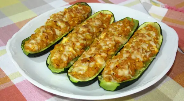 Cuatro calabacines rellenos de atún, gratinados con queso, servidos en un plato blanco sobre un mantel a cuadros.