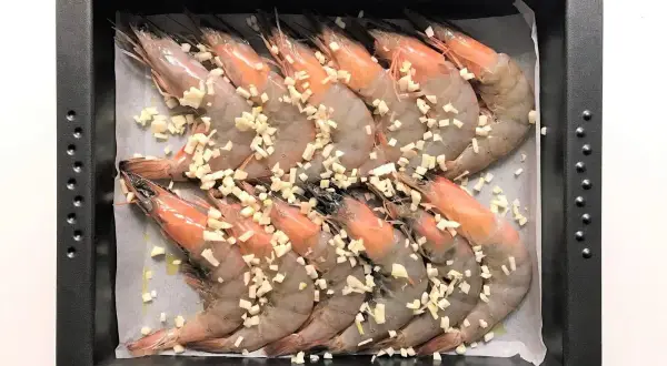Langostinos al horno listos para cocinar, cubiertos con ajo picado en una bandeja.