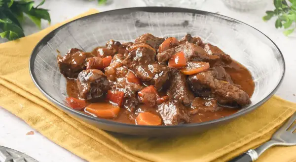 Plato humeante de carne al toro, tiernos trozos de carne en salsa oscura con zanahorias, servido en un cuenco gris sobre servilleta amarilla.