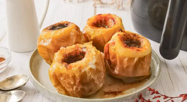 Cuatro manzanas asadas en freidora de aire, doradas y jugosas, servidas en un plato con un jarrito blanco al fondo.