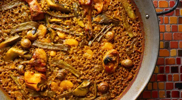 Paella de pollo con trozos de pollo dorado, judías verdes y caracoles, servida en una paellera sobre un mosaico de colores.